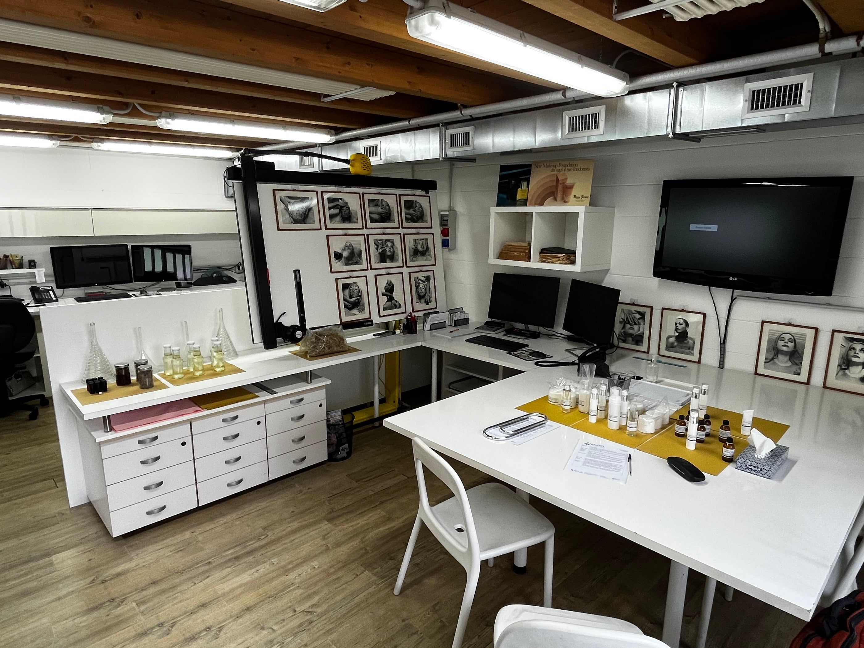 A well-organized art studio with white tables and chairs. It features computers, framed photos on walls, shelves with art supplies, and various bottles on tables. The ceiling has exposed wooden beams and visible ductwork, creating a modern industrial feel.