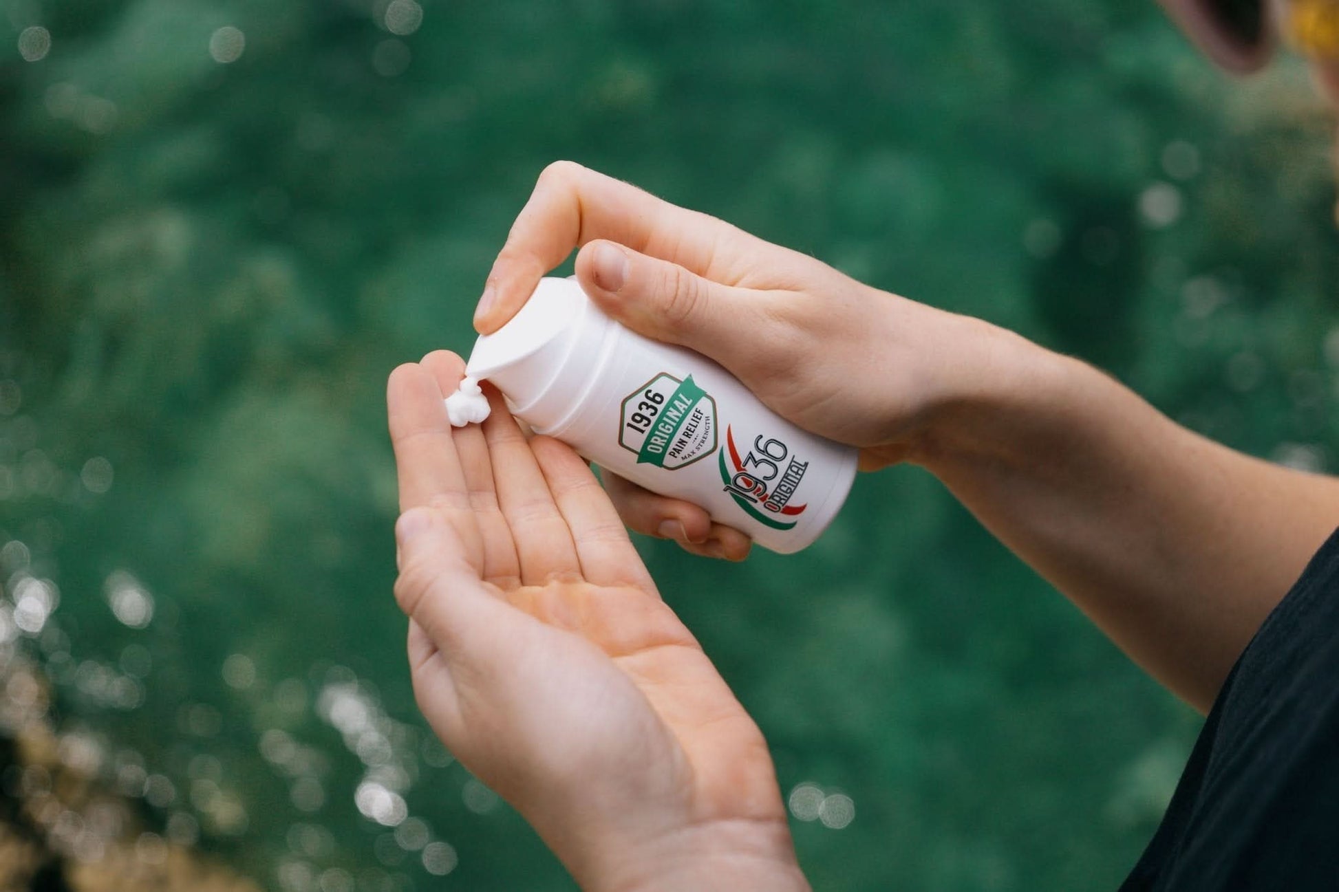 A person is dispensing a small amount of 1936 Original's Original Pain Cream, recognized for its natural formula, into their palm. The background features a blurred view of water, evoking a tranquil, natural setting.